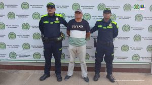 el capturado esta de frente custodiado por dos Policías. Detrás banner de la Policía Nacional.