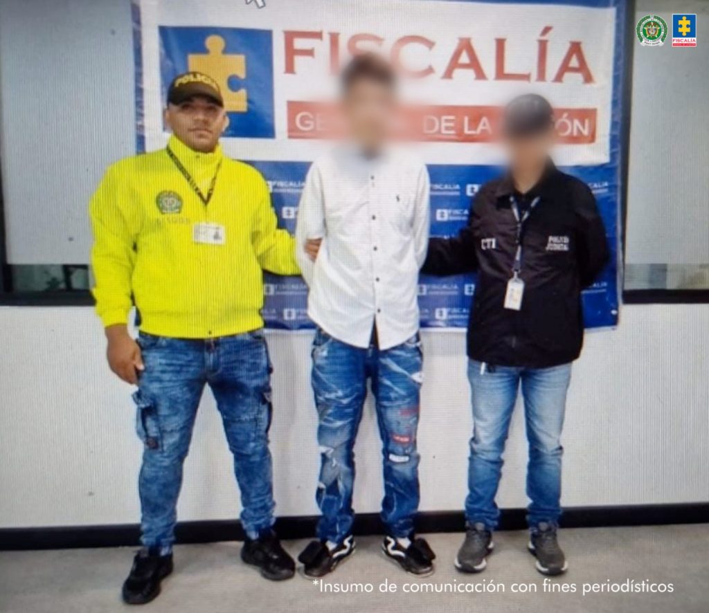 Se visualiza al capturado junto a un uniformado de la Policía Nacional y un investigador del CTI. Detrás de ellos el banner que identifica a la Fiscalía General de la Nación.