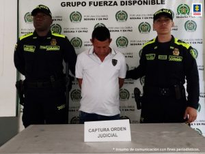 En la fotografía aparece un capturado, junto a personal de Policía Nacional. En la parte posterior de la imagen se ven banners de Policía Nacional y la Fiscalía General de la Nación.