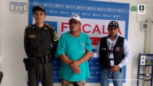 capturado esposado de frente, custodiado por investigador del CTI de la Fiscalía y uniformado de la Policía. Detrás banner de la Fiscalía.