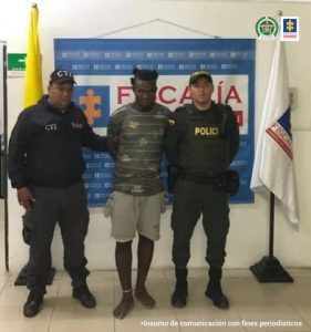 En la foto aparece de pie alias ‘Mano Biónica’, con los abrazos atrás; vestido con camiseta gris estampada, bermudas color gris y descalzo. Al lado derecho hay un servidor del CTI y a su izquierda un uniformado de la Policía Nacional. Detrás de ellos hay un pendón institucional de la Fiscalía y las banderas de Colombia y de la Fiscalía.