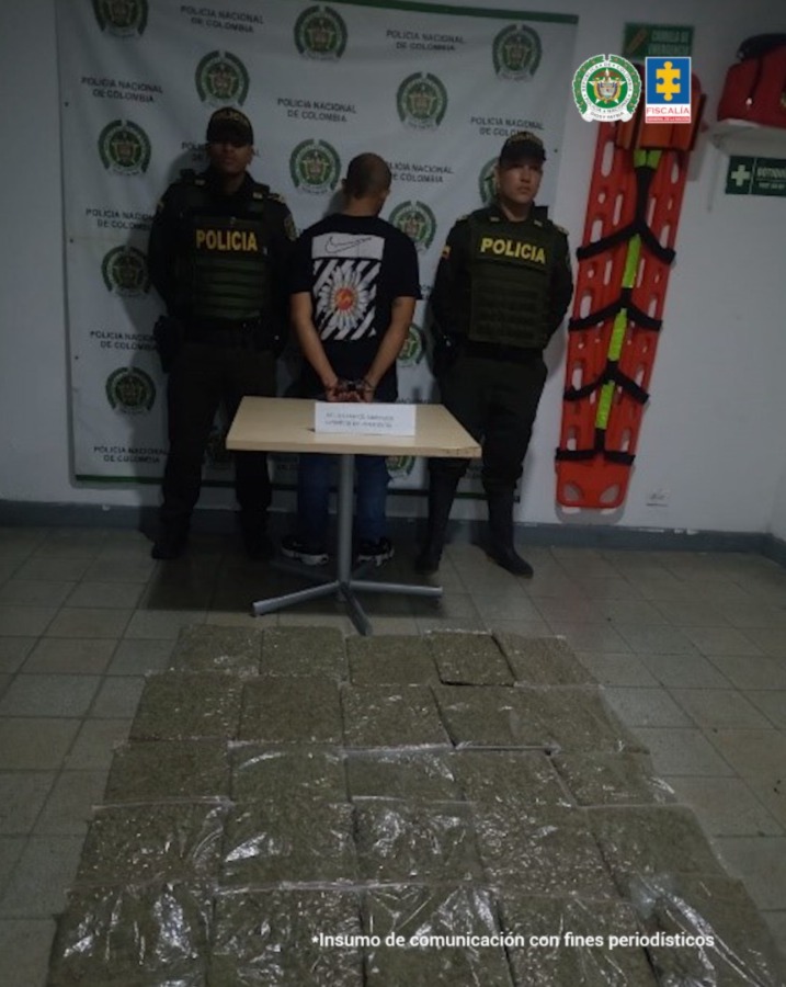 En la fotografía aparece de espaldas a la cámara, con los brazos atrás y esposado José Santiago Rendón Saldarriaga. Viste con camiseta de mangas cortas, de color negro y estampado grande en la espalde con colores blanco y rojo; pantalón oscuro. Delante hay una mesa y sobre ella hay un letrero con contenido borroso. En el piso se aprecian varios paquetes de plástico transparente con contenido vegetal. En la parte posterior, hay un pendón institucional de la Policía.