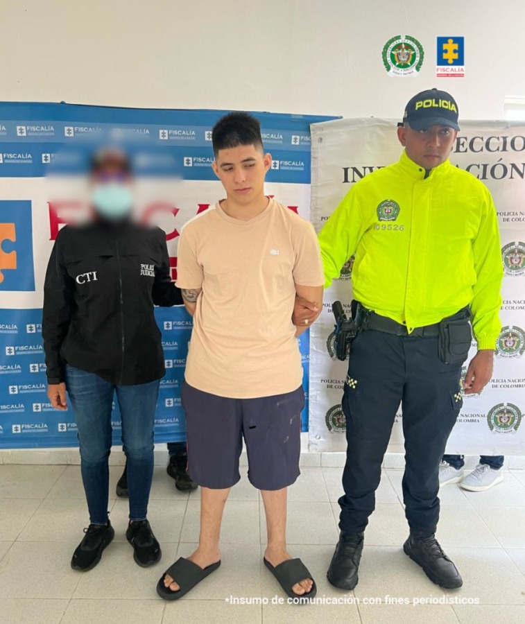 Se observa al capturado junto a una investigadora del CTI y un uniformado de la Policía Nacional. Detrás de ellos el banner que identifica a la Fiscalía General de la Nación y a la Policía Nacional.