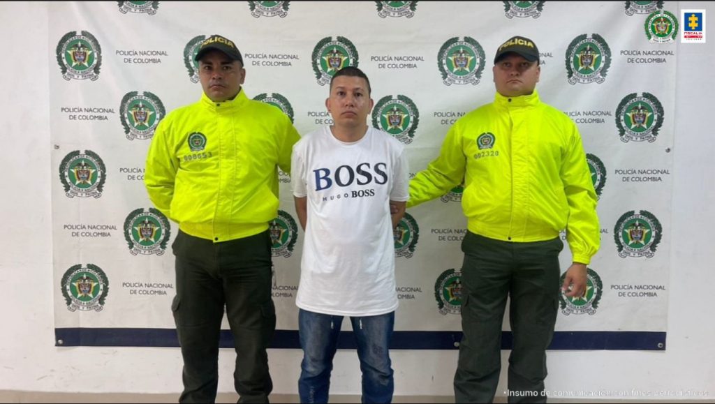 En la fotografía se aprecia de pie y con los brazos atrás a Juan Camilo Arango Villada. Viste camiseta de manga corta blanca y en la parte superior un estampado con la palabra ‘Boss’ en azul; jean azul y zapatillas. A lado y lado de él hay uniformados de la Policía Nacional. En la parte de atrás de ellos hay un pendón institucional de la Policía Nacional.