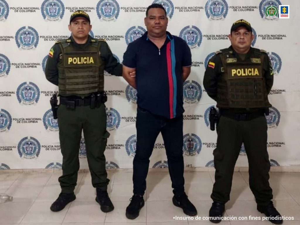 En la fotografía aparece un hombre capturado, acompañado de dos uniformados de la Policía Nacional. En la parte posterior un banner con logos de la entidad.