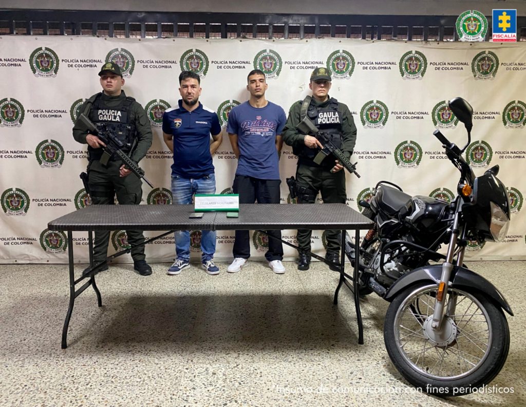 En la imagen están los dos procesados con un uniformado de la Policía Nacional a cada lado, además, una mesa con los 2 celulares incautados y la motocicleta.