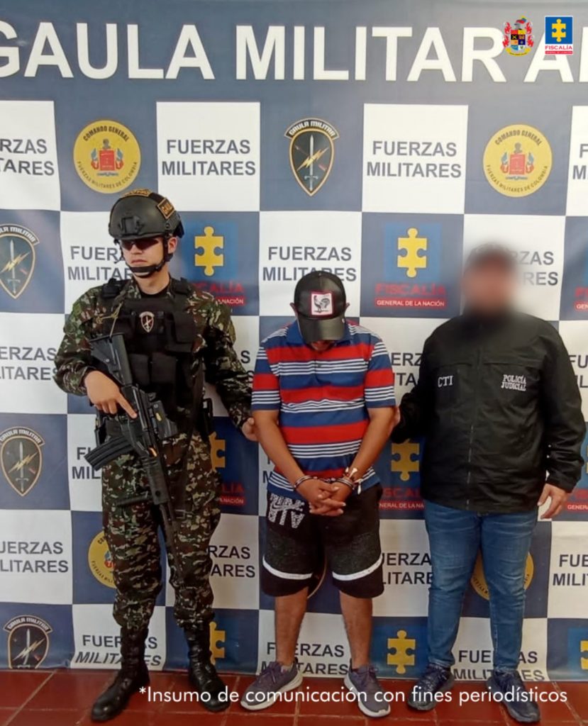 capturado de frente cabeza baja con gorra, custodiado por un investigador del CTI de la Fiscalía y un militar. Detrás banner del Grupo Gaula Ariari.