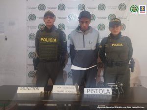 En la fotografía se aprecia, en acción de caminata, a Juan Carlos Sandoval Corrales. Viste sudadera azul con gris y gorra azul. A lado y lado de él lo custodian dos uniformados de la Policía Nacional. Delante de ellos hay una pesa y sobre ella hay un arma de fuego, un proveedor y munición. Asimismo, hay tres letreros que dicen: ‘Munición’, ‘Porte ilegal de armas de fuego’ y ‘Pistola’. Detrás de ellos hay un pendón institucional de la Policía.
