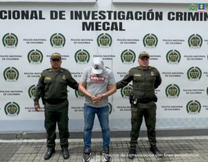 En la fotografía aparece de pie, cabeza inclinada hacia delante, con los brazos adelante y esposado Juan Carlos Serrano Segura. Viste con camiseta, de mangas cortas, de color gris, con estampado rojo y letras blancas a la altura del pecho; usa gorra de color blanco, jean azul desgastado y zapatillas negras. En los extremos del imputado hay uniformados de la Policía Nacional. Detrás de ellos hay un pendón institucional de la Policía.