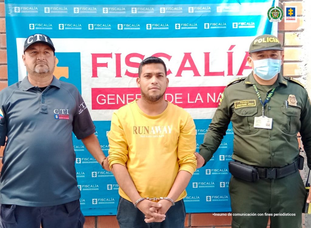 En la fotografía aparece un capturado, junto a personal de Fiscalía General de la Nación y Policía Nacional. En la parte posterior de la imagen se ven banners de Policía Nacional y la Fiscalía General de la Nación.    