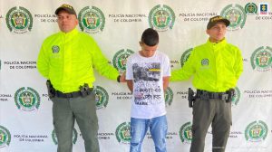 En la fotografía aparece un capturado junto a personal de Policía Nacional y Fiscalía General de la Nación. En la parte posterior de la imagen se ven banners de Policía Nacional y la Fiscalía General de la Nación.