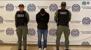  Se observa un capturado de frente. A cada costado un uniformado de Policía Nacional y atrás un pendón de Policía Nacional 