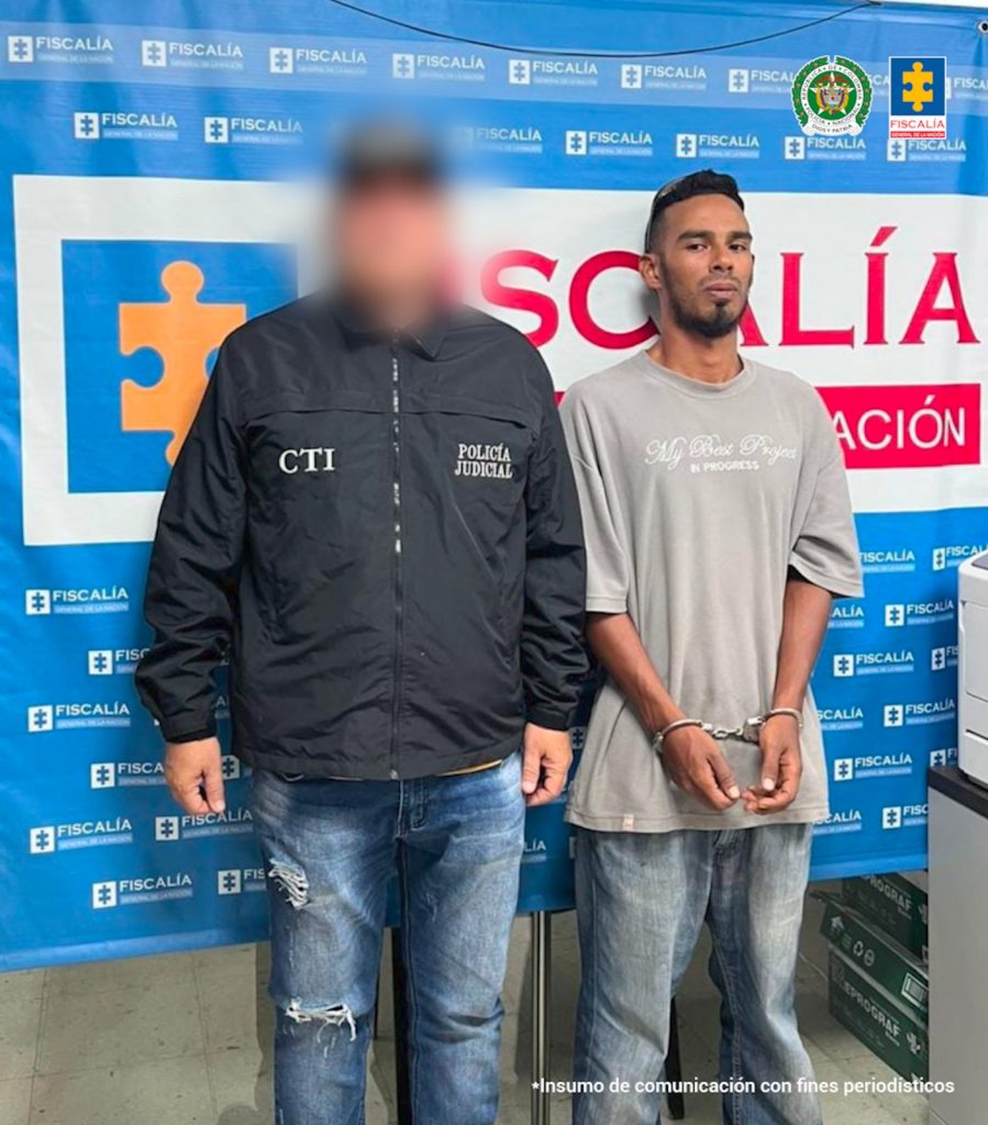 En la fotografía aparece de pie, con los brazos adelante y esposado Juan David Pino Cardona. Viste con camiseta de color gris, mangas cortas y estampado blanco a la altura del pecho; jean azul claro. En su extremo derecho hay un servidor del CTI de la Fiscalía. Detrás de ellos hay un pendón institucional de la Fiscalía.