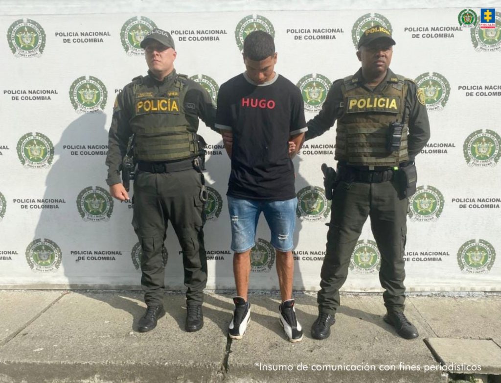 En la fotografía aparece de pie, la cabeza inclinada hacia adelante y con los brazos atrás Juan David Rodríguez. Viste con camiseta de mangas cortas, de color negro y con letras de color rojo estampadas a la altura del pecho; bermuda de jean azul desgastadas y zaptillas negras con blanco. En sus extremos hay uniformados de la Policía Nacional. Detrás de ellos hay un pendón institucional de la Policía.