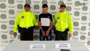 En la fotografía aparece el procesado con 2 servidores de la Policía Nacional y en una mesa el celular incautado