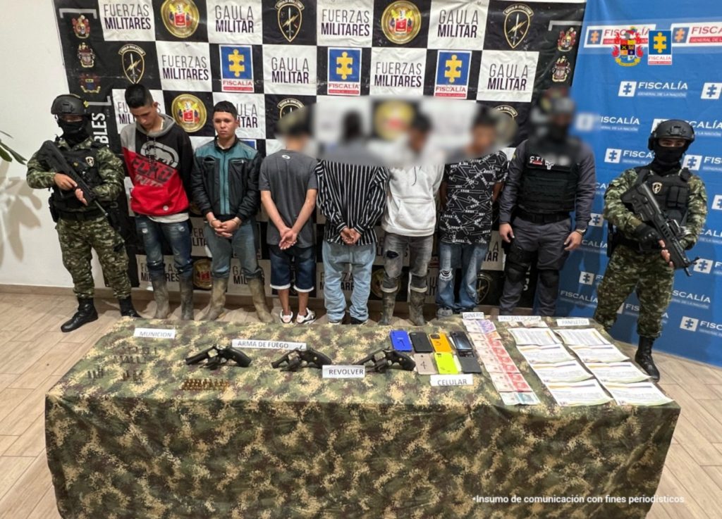 En la fotografía, aparecen de pie Juan Diego Alfonso Montes, alias Juan Diablo; y Luis Miguel Peña Gutiérrez, alias Kevin. Uno viste buso con los colores negro, rojo y gris; jean azul desgastado y botas pantaneras. El otro, con los brazos adelante y asposado, usa chaqueta de colores negro y verde; jean azul y botas pantaneras. A su izquierda hay un servidor del CTI de la Fiscalía y un uniformado del Ejército. En el extremo derecho hay un uniformado del Ejército. Delante de ellos hay una mesa con mantel camuflado y sobre ella se ve munición, dos armas de fuego, celulares, dinero en efectivo y panfletos. Detrás de ellos hay dos pendones institucionales de la Fiscalía y otro con logos de Fiscalía y del Ejército.