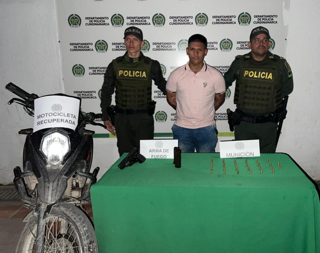 fotografia en donde se ve al capturado en medios de dos policias, delante de ellos se encuentra una mesa con el material incautado, al lado de ella uan moticicleta hurtada, detras de ellos hya un baking de la policia