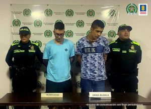 En la foto se aprecian de pie y con los brazos atrás a Juan Felipe Álvarez Muñoz y Jhoan Sebastián Solarte Muñoz. Uno viste camiseta de manga corta azul clara, pantaloneta negra, usa lentes y cabello corto rapado a los lados. El otro viste camiseta de manga corta con estampado blanco y azul, franja azul a la altura del pecho con las palabras ‘Blue’ y ‘Ing’. A lado y lado de ellos hay uniformados de la Policía Nacional. Delante de ellos hay una mesa marrón y sobre ella hay se aprecia un cuchillo y dos letreros que dicen: ‘Homicidio’ y ‘Arma blanca’. Detrás de ellos hay un pendón institucional de la Policía.