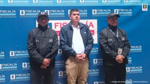 En la fotografía se encuentra el procesado con una chaqueta azul y servidores del CTI  a cada lado. Al fondo hay un pendón de la Fiscalía General de la Nación azul con un mosaico del logo de color blanco y en el centro un logo de tamaño grande de la misma entidad 