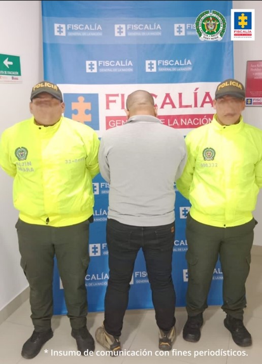 Se visualiza al capturado, junto a dos uniformados de la Policía Nacional. Detrás se ubica el banner que identifica a la Fiscalía General de la Nación.