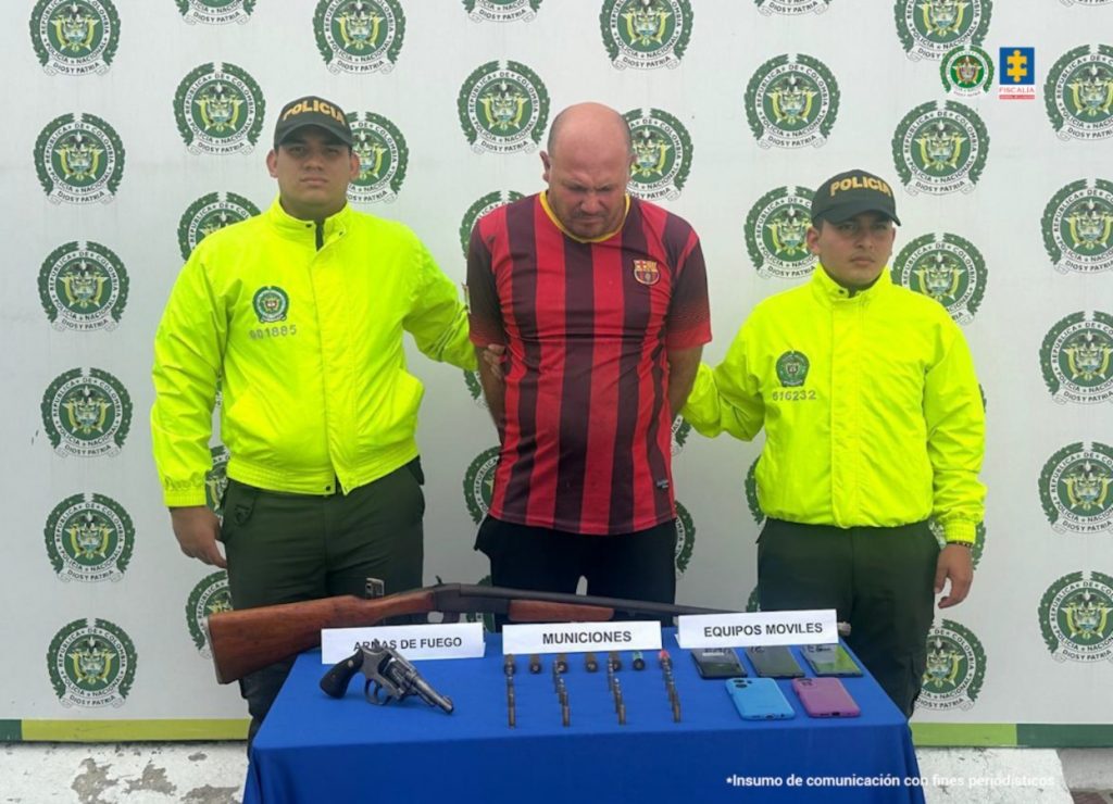 Se visualiza al capturado junto a dos uniformados de la Policía Nacional. Frente a ellos una mesa con las armas, munición y teléfonos incautados. Detrás el banner que identifica a la Policía Nacional.
