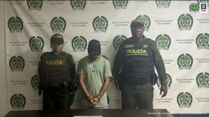 En la fotografía aparece de pie, la cabeza agachada, los brazos adelante y esposado Juan José Torres Marín. Viste con camiseta verde clara de manga corta y gorra negra. A lado y lado de él hay uniformados de la Policía Nacional. Delante de ellos hay una superficie color marrón y sobre ella hay un letrero que dice: ‘Violencia intrafamiliar’. Detrás de ellos hay un pendón institucional de la Policía.