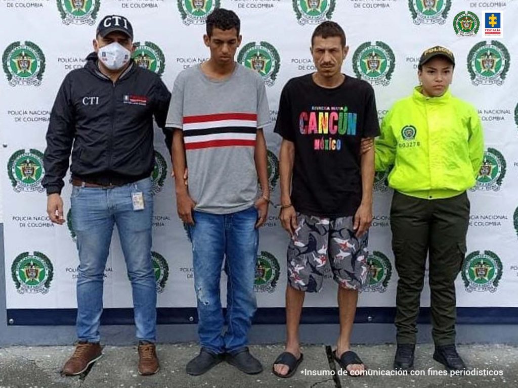 En la foto aparecen de pie, Pedro Antonio Muñoz Marín, alias ‘Torrino’ y Juan José Uribe Reyes, alias ‘Muelas’. Uno de los imputados vista camiseta gris de manga corta con franjas horizontales de color rojo, blanco, azul; jean azul y zapatos negros. La otra persona viste camiseta de manga corta negra con estampado a la altura del pecho, pantaloneta estampada y pantuflas oscuras. Al lado derecho de ellos hay un servidor del CTI de la Fiscalía y a la izquierda una uniformada de la Policía Nacional. Detrás de ellos hay un pendón institucional de la Policía.