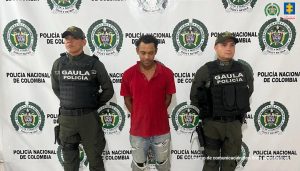 En la fotografía aparece una persona capturada en compañía de uniformados de la Policía Nacional. En la parte posterior un banner con logos de la entidad. 