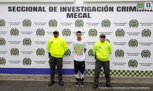 En la fotografía se aprecia a Juan Manuel Galindo Lenis, de pie y con los brazos atrás. Viste camiseta de manga corta blanca con estampado de colores verde y naranja; pantalón de sudadera negro con blanco y zapatillas negras con blanco. A lado y lado de ellos hay uniformados de la Policía Nacional y Detrás de ellos hay un pendón institucional de la Policía.