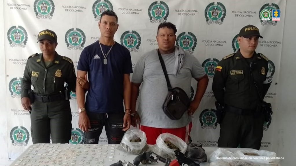 En la fotografía aparecen de pie y esposados entre sí Juan Manuel Llano Rendón y Jhon Stic Mazuera Acevedo. Uno viste con camiseta de mangas cortas de color azul oscuro y pantalón negro con estampados blancos y rojos. Usa reloj en la muñeca derecha; así como dos manillas en la muñeca izquierda. También usaccadena de plata con dije en forma de placa en el cuello. El otro, viste con camiseta de mangas cortas, de color gris y pantalón de color rojo, con franjas blancas a los lados y usa maletín de color negro atravesado, así como una tela en su hombro izquierdo. En los extremos de ellos hay uniformados de la Policía Nacional. Delante de ellos hay una superficie blanca y sobre ella se aprecian gran cantidad de papeletas de plástico transparente con contenido blanco o beage; tres armas de fuego, munición, así como bolsas y paquetes de plástico transparente con contenido vegetal. En la parte de atrás hay un pendón institucional de la Policía.