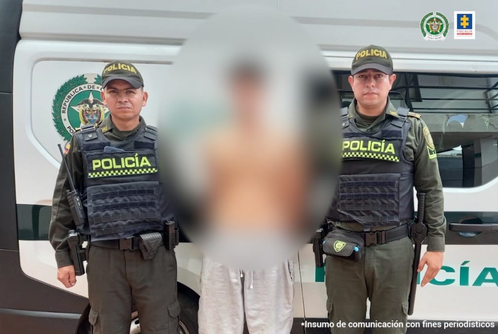 capturado en pantaloneta sin camisa esposado, custodiado por dos policías, detrás patrulla de la Policía.