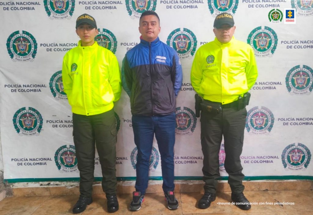 Se observa un hombre vestido con chaqueta en azul y negro de frente y a cada costado un funcionario de Policía Nacional. Al fondo un pendón de Policía Nacional