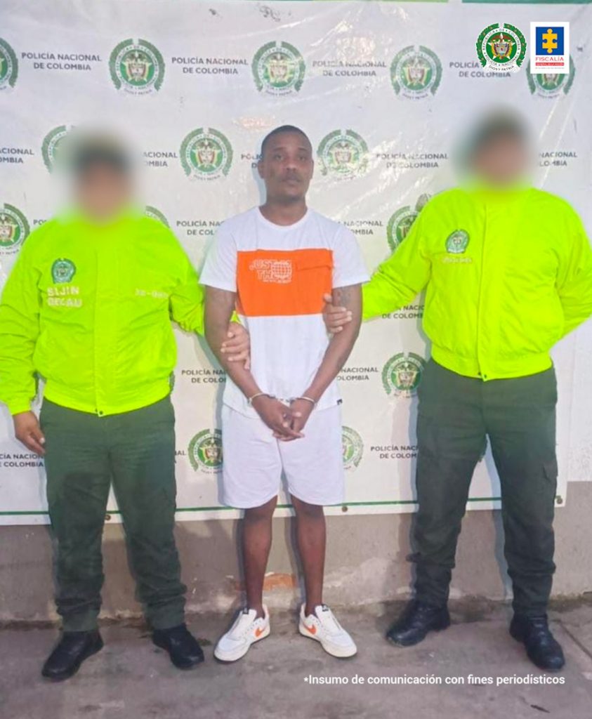 En la foto aparece, de pie, con los brazos adelante y esposado, Juan Sebastián Cabal Martínez, alias ‘Cabal’. Viste camiseta de manga corta blanca con una franja color naranja a la altura del pecho; pantaloneta y tenis blancos. A lado y lado de él hay uniformados de la Policía Nacional. Detrás de ellos hay un pendón institucional de la Policía.