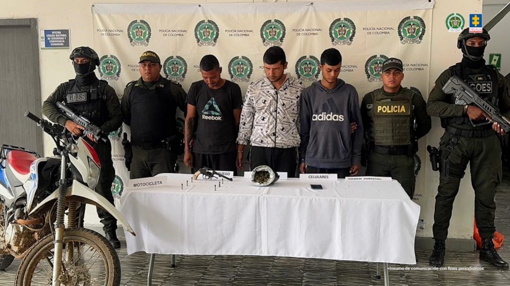 En la fotografía aparecen en hilera, de pie y esposados entre sí Juan Sebastián Ramírez Giraldo y Cristian Stiben Pérez Gómez y Víctor Alfonso Cañas Zuleta. Uno viste camiseta de color negro, de manga corta y estampado en forma de triángulo en pecho; así como con pantalón negro. Otro viste con camisa de manga larga, d color blanco con estampados irregulares de color azul y dorado; y pantalón negro. El otro viste con buso de color gris, de manga larga, con estampado blanco en el pecho y pantalón negro. En los extremos de ellos hay uniformados de la Policía Nacional. Delante de ellos hay una mesa con mantel blanco y sobre ella hay un arma de fuego, munición, un celular y una bolsa. En el extremo izquierdo de la mesa hay una motocicleta. Detrás de ellos hay un pendón institucional de la Policía.