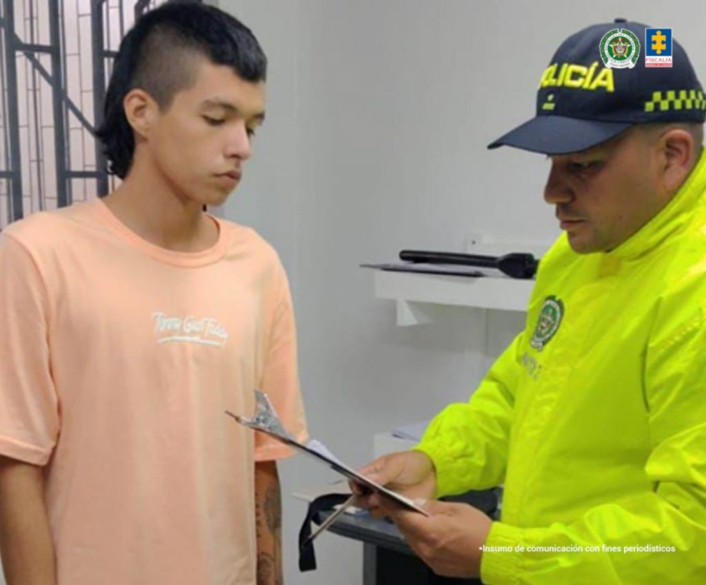 En la fotografía tomada en interiores, se aprecia de pie a Juan Sebastián López Mejía. Viste camiseta de manga corta y de color salmó, con estampdo blanco en el pecho. Frente a él hay un uniformado de la Policía Nacional en acción de lectura de unos documentos que tiene en sus manos.