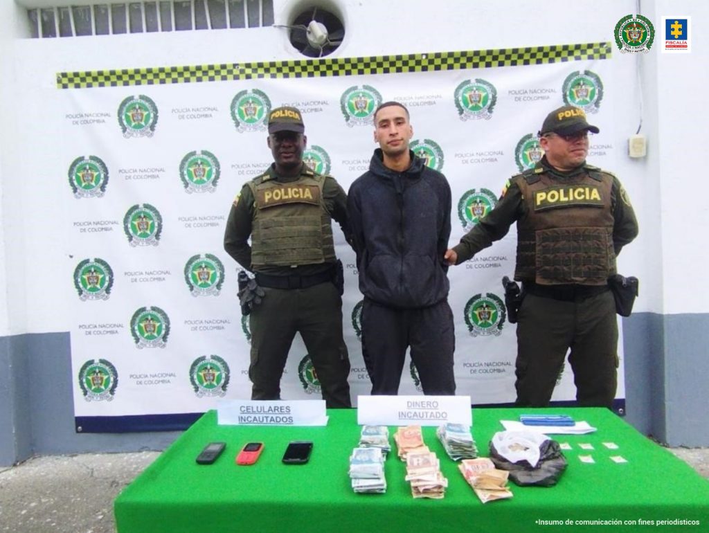 En la foto aparece de pie, con los brazos atrás, Juan Sebastián Quiroga Caicedo. Viste buzo y pantalón oscuro. Al lado y lado de él hay uniformados de la Policía Nacional. Delante de ellos hay una mesa con superficie verde y sobre ella se ven tres celulares, dinero en efectivo, una bolsa negra y varias papeletas. Asimismo, hay dos letreros que dicen: ‘celulares incautados’ y ‘dinero incautado’. Detrás de ellos hay un pendón interinstitucional de la Policía.