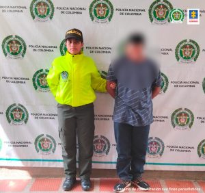 capturado de frente esposado manos atrás custodiado por policías. Detrás banner de la Policía Nacional.