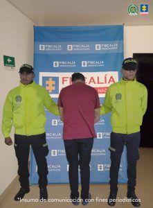 Se visualiza al capturado de espaldas junto a dos uniformados de la Policía Nacional. Detrás de ellos el banner que identifica a la Fiscalía General de la Nación.
