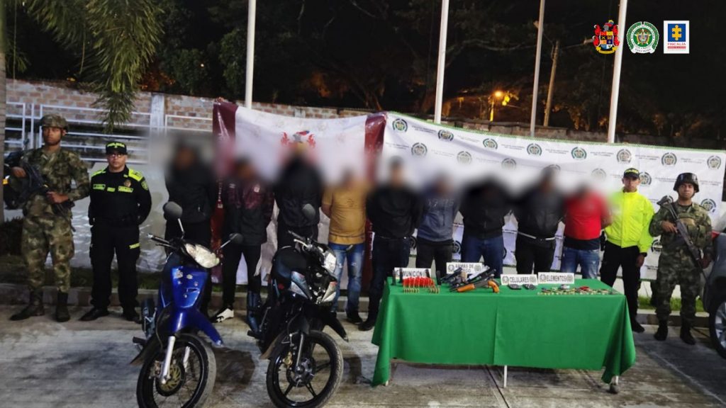 En la fotografía se aprecia a los hoy imputados con la cabeza agachada y los brazos atrás. A los costados de ellos hay uniformados de la Policía Nacional y del Ejército. Delante de ellos hay una mesa con mantel verde y sobre ella varios elementos como munición, armas de fuego, celulares, entre otros. Asimismo, al lado derecho se aprecian dos motocicletas y al lado izquierdo un vehículo gris. En la parte posterior hay dos pendones institucionales del Ejército y de la Policía.