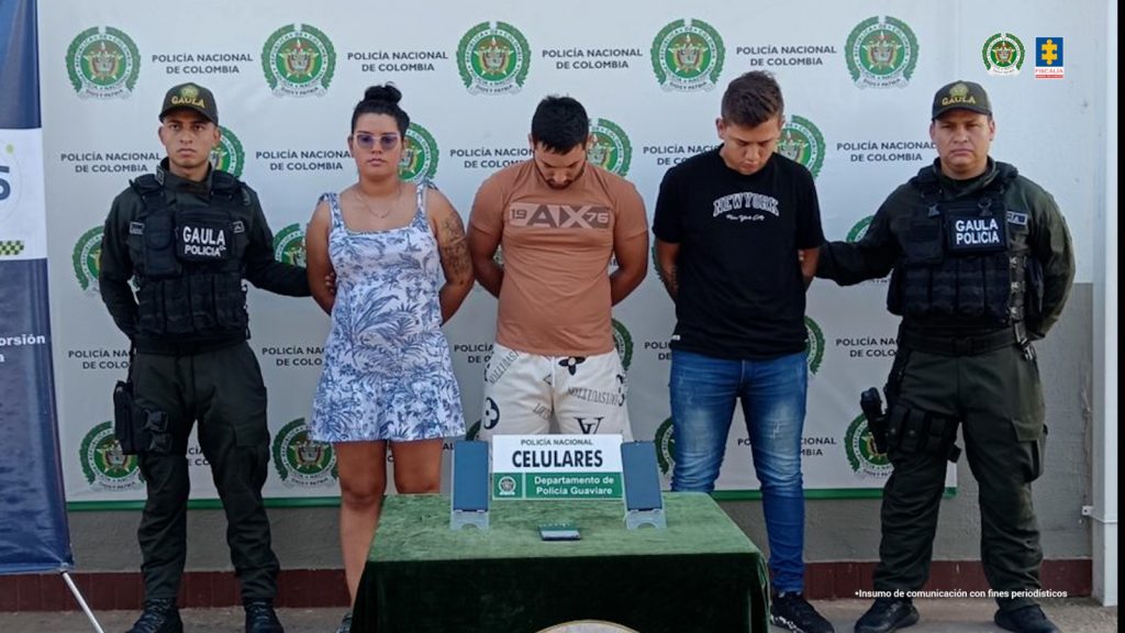 capturados de frente custodiados por dos policías, delante de ellos una mesa con material incautado. Detrás banner de la Policía Nacional.