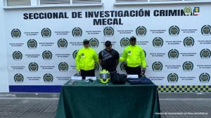 En la fotografía se aprecia de pie y con los brazos atrás a Julio César Hoyos Vaca. Viste con camiseta negra de manga corta, con estampado blanco en la parte superior derecha. A sus extremos hay uniformados de la Policía Nacional. Delante de ellos hay una mesa con mantel verde y sobre ella hay dinero en efectivo, un arma de fuego, un casco de motociclista verde, una maleta y otros elementos de color negro. Detrás de ellos hay un pendón institucional de la Policía.