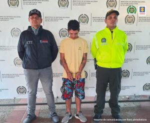  capturado de frente esposado custodiado por un investigador del CTI de la Fiscalía y un Policía. Detrás banner de la Policía Nacional.