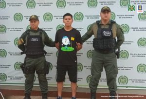 capturado de frente custodiado por dos policías. Detrás banner de la Policía Nacional.