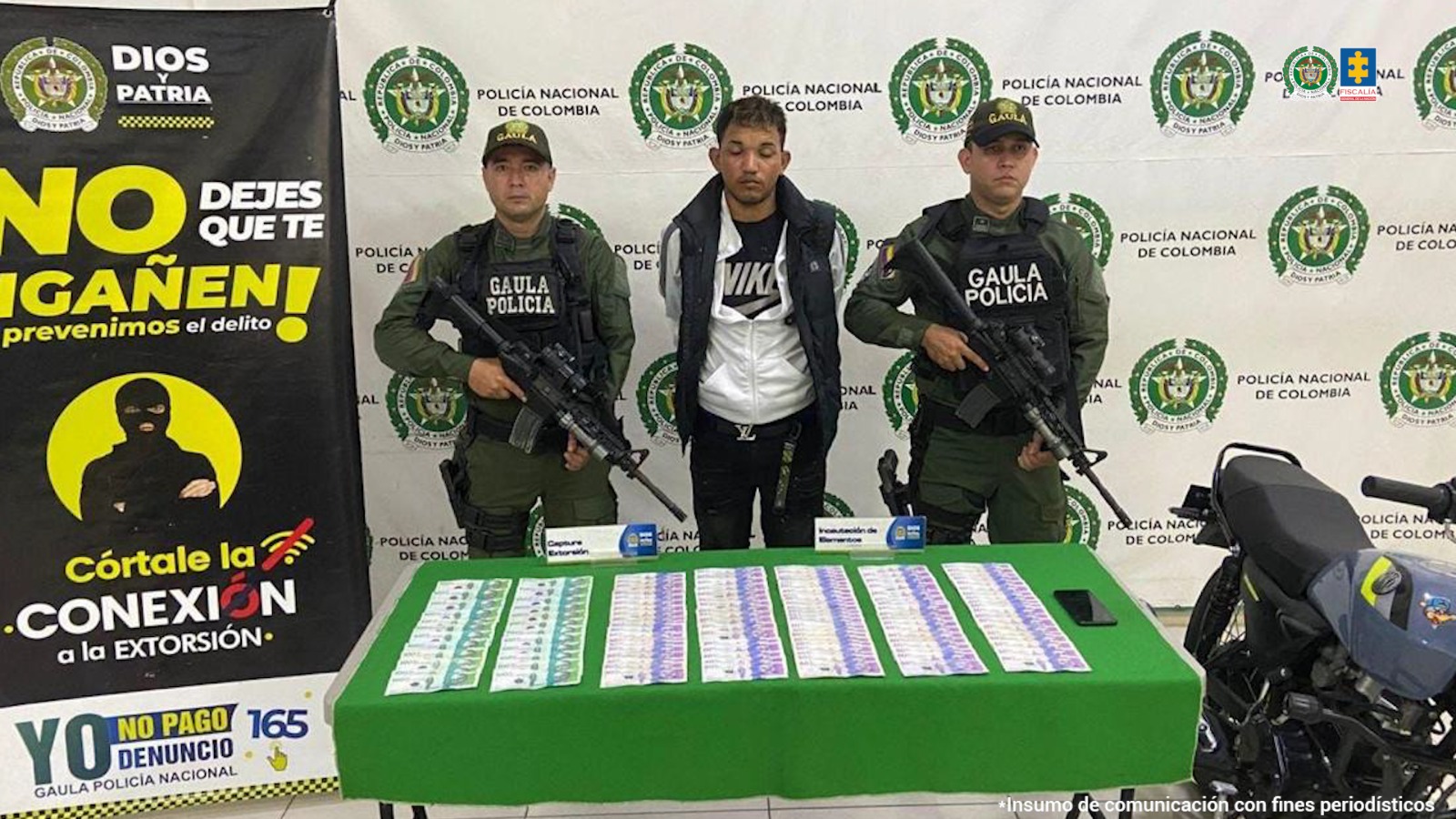 El capturado aparece detrás de una mesa en la que hay billetes de diferentes denominaciones. Dos personas del Gaula rodean al capturado. Al fondo se ve un banner de la Policía. 