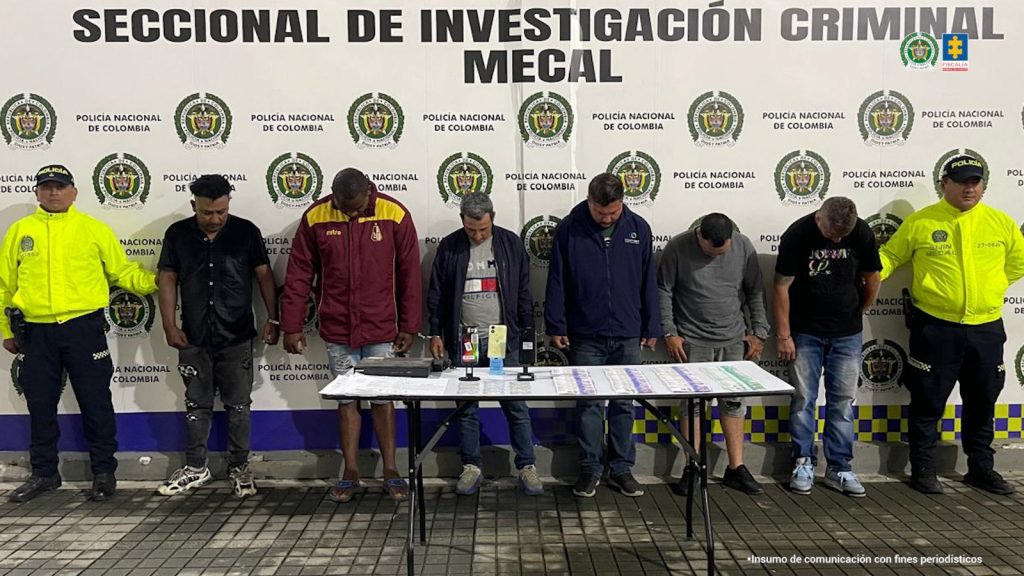 En la Fotografía aparecen de pie, esposados entre sí y con la cabeza agachada los seis presuntos implicados en hurto de vehículos. A sus extremos hay uniformados de la Policía Nacional. Delante de ellos hay una mesa y sobre ella se aprecian celulares, dinero en efectivo y documentos. Detrás de ellos hay un pendón institucional de la Policía.