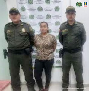 En la fotografía aparece una mujer capturada, acompañada de dos uniformados de la Policía. En la parte posterior un banner con logos de la entidad.