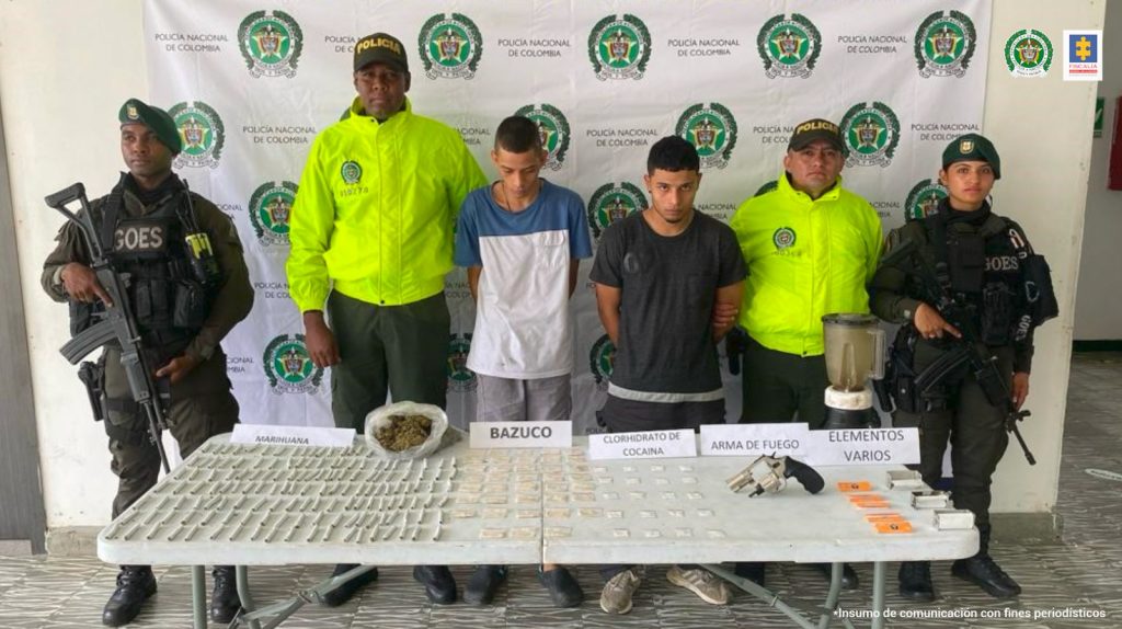 En la fotografía se aprecia a dos de los tres presuntos implicados en el hurto a la fundación de seguidores de un equipo de fútbol. Uno viste buzo de sudadera con franjas azul, blanca y negra y la marca ‘Puma’ en el torso; pantaloneta camuflada y pantuflas. La otra persona viste camisa de la Selección Colombia azul; jean con desgaste y zapatillas. A lado y lado de ellos hay uniformados de la Policía Nacional y detrás de ellos hay un pendón institucional de la Policía.