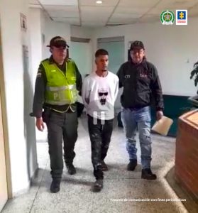 En la foto aparece en un pasillo, en acción de desplazamiento, con los brazos atrás, Kevin Alexis Ramírez Valencia. Viste buzo blanco con estampado en el centro, pantalón y zapatos oscuros. Al lado derecho de él hay un uniformado de la Policía Nacional y al lado izquierdo un servidor del CTI de la Fiscalía.