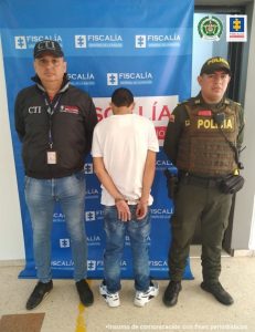 está de espaldas esposado manos atrás, custodiado por investigador CTI de Fiscalía y Policía. Detrás banner de la Fiscalía.