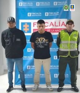 En la imagen se ve un hombre detenido en custodia de un investigador del CTI y un integrante de la Policía Nacional. Detrás suyo un backing de la Fiscalía.